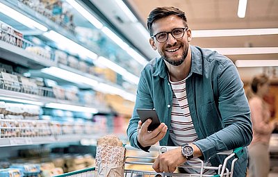 Young man with beard and glasses happily leaning on shopping cart in supermarket with his smartphone in hand