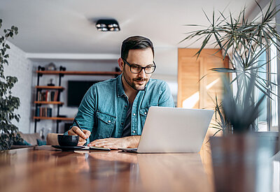 Man works on a notebook in his home office and writes down notes on a piece of paper