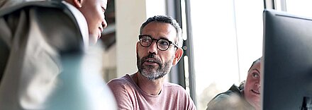 Middle-aged man with gray hair, beard and glasses wearing pink shirt listens intently to his younger dark-skinned female colleague while working at a PC in an office