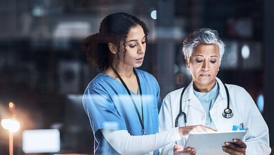 Young nurse with black curly hair and blue nurse's coat shows something on a tablet to an older senior doctor with short gray hair in a white doctor's coat and a stethoscope around her neck