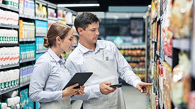 A supermarket employee with a tablet checks the goods on the shelf with her colleague with a handheld scanner