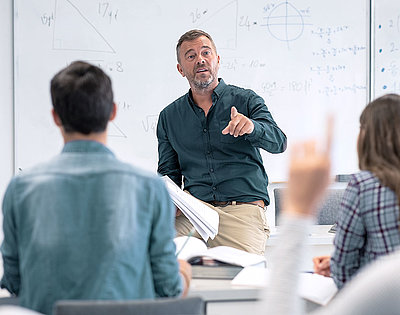 LANCOM requirements for a good school Wi-Fi Middle-aged teacher stands in front of whiteboard with mathematical formulas and calls on student who is about to report in
