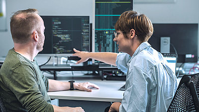 An IT specialist and an IT specialist consult on IT measures in front of several monitors with programming language