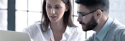 Brunette young woman in white shirt explains something on notebook in light office to her brunette colleague of the same age with beard, glasses and light blue shirt