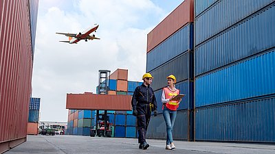 Photo: Two people wearing safety helmets walk between tall stacks of containers at a container terminal; in the background, a container handler is operating, and a passenger aircraft is flying overhead.