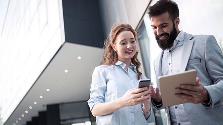 Photo: Two people standing in front of a modern building while jointly looking at a smartphone and tablet.
