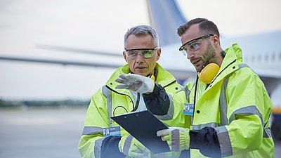 Photo: Two employees wearing yellow protective clothing and hearing protection stand in front of an aircraft and discuss documents on a clipboard.