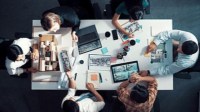 Photo: Several people sitting at a table with architectural models, floor plans, tablets, and a laptop while collaboratively planning a building system.
