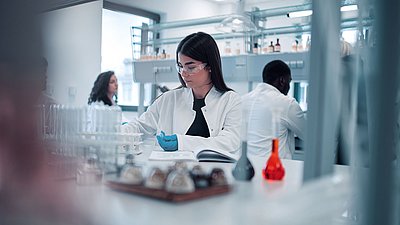 Photo: Several people working in a modern laboratory; in the foreground, a woman wearing safety glasses and gloves is seated at a laboratory bench with glass containers and samples.