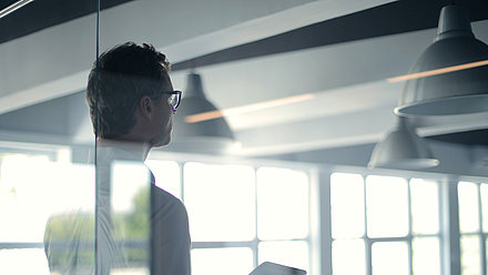 Man standing behind glass wall of office with tablet in hand