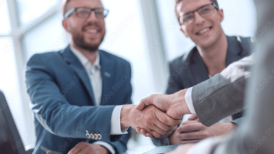 Photo of a handshake between two people in suits, with other colleagues in the background at a business meeting in a modern office.