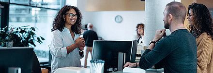 Administrative employee with black hair and glasses greets friendly young couple at her digital workplace