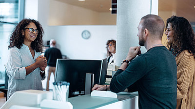 Administrative employee with black hair and glasses greets friendly young couple at her digital workplace