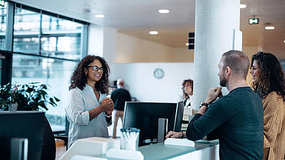 Administrative employee with black hair and glasses greets friendly young couple at her digital workplace