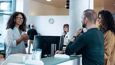 Administrative employee with black hair and glasses greets friendly young couple at her digital workplace