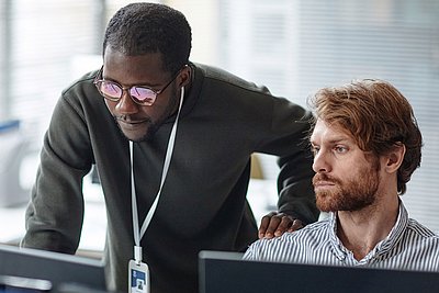 Two coworkers focus on computer screens, one standing and guiding the other in a modern office setting.