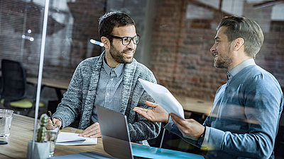 Two male colleagues talking happily at desk in office