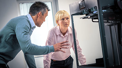 Blonde middle-aged woman with short hair animatedly discussing with her male colleague in front of a machine