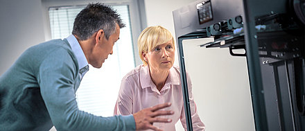 Blonde middle-aged woman with short hair animatedly discussing with her male colleague in front of a machine