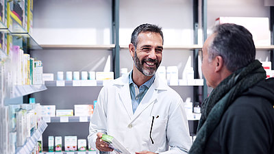 Two men in a pharmacy; Pharmacist with short black hair and beard; wears a doctor's coat and holds a medicine in his hand. He smiles at man standing in front of him, also black short hair, jacket and scarf.