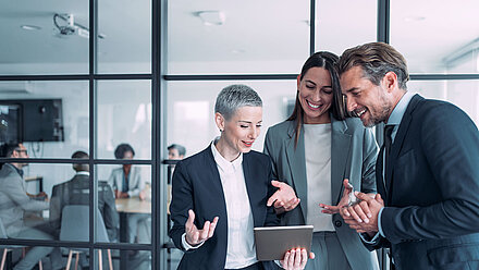 A short-haired, neat woman in a blazer with gray hair (left) discusses a successful project with a brunette, long-haired colleague in a gray blazer holding a tablet and a young colleague with curly dark blue hair in business attire in the right half of the picture; in the background, other office employees can be seen through a glass front in a meeting room