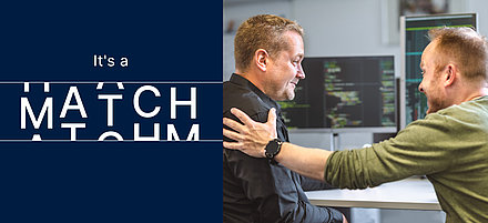 Two-part advertising banner: On the left is the white text "It's a Match" on a dark blue background; on the right are two middle-aged gentlemen sitting in front of PC monitors showing programming language. One taps the other confidently on the shoulder and smiles