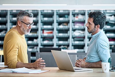 Older gentleman with glasses, gray hair and yellow sweater stands with younger colleague with black hair and shirt in front of individual parts store and discusses on laptop the digititalization of their business