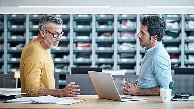 Older gentleman with glasses, gray hair and yellow sweater stands with younger colleague with black hair and shirt in front of individual parts store and discusses on laptop the digititalization of their business