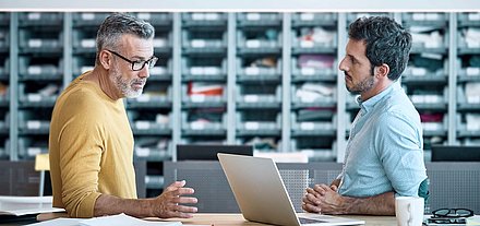 Older gentleman with glasses, gray hair and yellow sweater stands with younger colleague with black hair and shirt in front of individual parts store and discusses on laptop the digititalization of their business