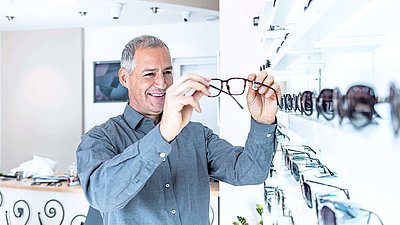 Older man with gray hair stands in front of the glasses shelf at an optician's and looks contentedly at a pair of glasses