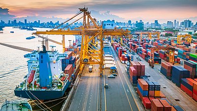 Photo: Large container port with cargo ship, cranes, and numerous container stacks in front of a city skyline.
