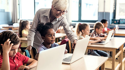 Blonde teacher with glasses checks digital task solved by elementary school student on laptop while every child in classroom works on laptop