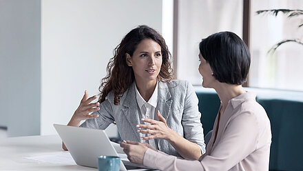 Two colleagues are sitting at a table in front of a laptop, one of them is gesturing with her hands.