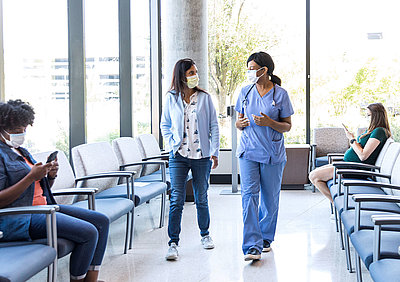 Young doctor in light blue jumpsuit picks up patient with mouth guard from bright waiting area of a clinic for her treatment, where a pregnant woman and another woman are waiting to be called up