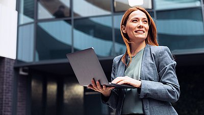 Photo: Woman with a laptop standing in front of a modern office building with a glass facade.