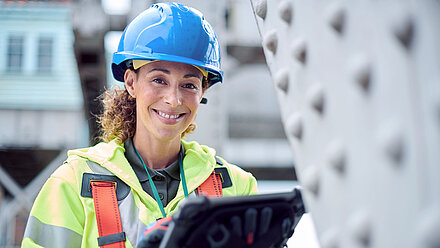 Woman with helmet and safety jacket holds tablet and smiles at camera