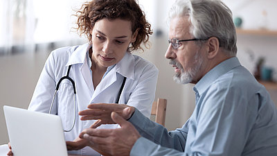 Two people are sitting at a table; Middle-aged female doctor (left) has short brown hair, wears a doctor's coat and a stethoscope around her neck. She is showing something on a laptop to an older man (right). The man has short gray hair, a gray beard, and glasses. He is wearing a blue shirt.