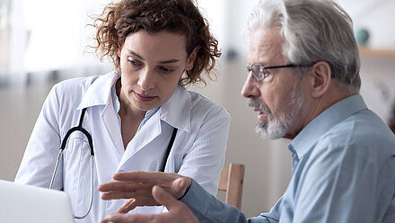 Two people are sitting at a table; Middle-aged female doctor (left) has short brown hair, wears a doctor's coat and a stethoscope around her neck. She is showing something on a laptop to an older man (right). The man has short gray hair, a gray beard, and glasses. He is wearing a blue shirt.