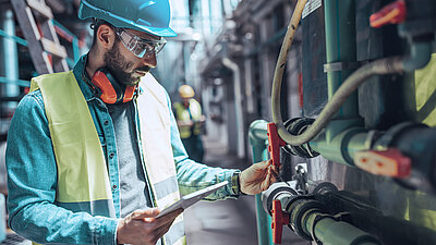 Photo of a plumber wearing goggles, a high-visibility vest, noise-canceling headphones and a hard hat checking pipes and valves in a supply system with a clipboard
