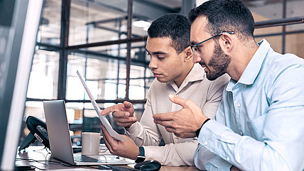 IT expert with beard and glasses explains a document to young IT employee in modern office