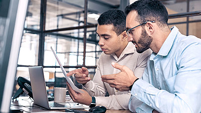 IT expert with beard and glasses explains a document to young IT employee in modern office