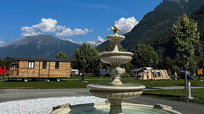 Stone fountain in foreground, wooden mobile home, tents, and mountains under blue sky on a campsite in background.