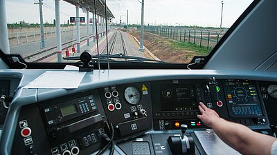 Photo: View from a train driver’s cab toward railway tracks and a station platform; control elements and displays are visible in the foreground.