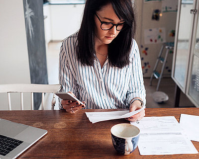 Young brunette woman with glasses sits at home at dining table with kitchen in background and tries to make a bureaucratic application with laptop, printouts, and cell phone