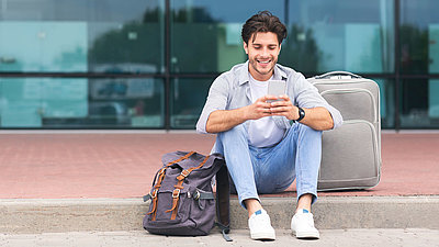 Traveling man sits with SMartphone and suitcase on sidewalk in front of airport terminal