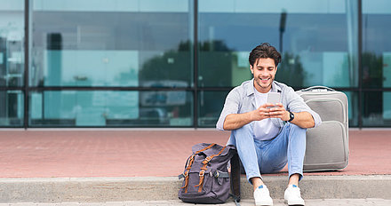 Traveling man sits with smartphone and suitcase on sidewalk in front of airport terminal