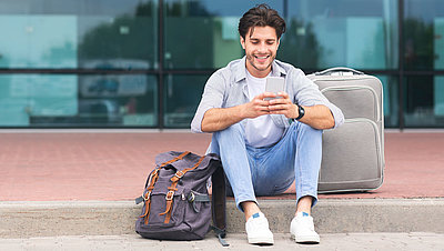 Traveling man sits with smartphone and suitcase on sidewalk in front of airport terminal