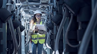 Photo: Female technician wearing a safety helmet, safety glasses, and a high-visibility vest standing among technical equipment and cable systems while holding a clipboard to inspect infrastructure components.