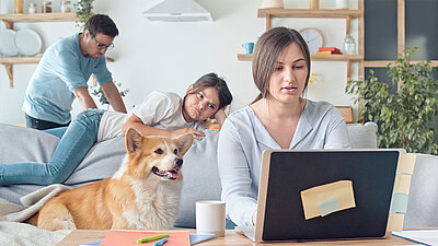 Woman working in home office on laptop with child, dog and husband in the background