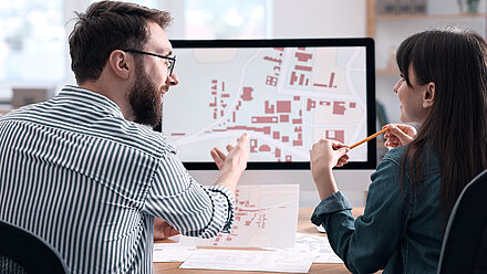 Photograph of two civil servants (brunette man with glasses, beard and striped shirt on the left and long-haired brunette woman with pencil and green blouse on the right) from behind, looking together at building location plans of a city at a desk